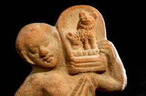 Römerzeitliche Terracotta-Figur aus Ägypten von einem jungen Mann, der auf seiner Schulter einen gepolsterten Korbsessel trägt, in dem ein kleiner spitzartiger Hund sitzt